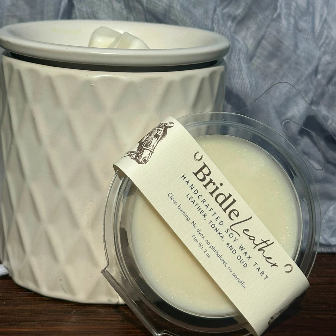 A close-up of a wax melt product labeled 'Brizzle Leather' with a clear plastic clamshell container in the foreground and a larger textured cream-colored wax vessel behind it on a dark wooden surface against a blue fabric backdrop.